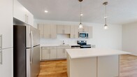 This thoughtfully designed single-floor unit offers a clean, modern aesthetic throughout its four rooms. The kitchen serves as the centerpiece of the space, showcasing flat-front cabinetry in a warm greige tone, a suite of stainless steel appliances including a refrigerator, electric range, and built-in microwave, and a white quartz countertop island with elegant pendant lighting overhead. Light hardwood flooring runs throughout the kitchen and living areas, lending warmth and continuity to the open layout. The bedroom areas are finished with plush, neutral-toned carpeting, creating a quiet and comfortable retreat. Each bedroom features clean white walls and window blinds that allow for natural light while maintaining privacy. The bathroom is well-proportioned and stylishly finished, featuring a wide vanity with white quartz countertops, an undermount sink, ample mirror space, hardwood-style flooring, and a walk-in shower with tiled walls. A walk-in closet with built-in shelving adds practical storage. Recessed lighting throughout provides consistent, modern illumination. The overall ambiance is bright, clean, and contemporary, making efficient use of the available footprint with a cohesive, minimalist design palette.