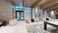 Located at 65 Broadway Ave in Toronto's Yonge and Eglinton neighbourhood, this generously proportioned amenity lounge offers a refined and contemporary communal experience across a single expansive floor. The space is defined by its striking architectural detailing, most notably the warm wood-panelled ceilings that run throughout, creating a cohesive and inviting atmosphere against crisp white walls and light-toned tile flooring.

Upon entry, a welcoming reception corridor leads into multiple distinct yet open zones. A bright lounge area near the building entrance features plush cream armchairs arranged around a textured area rug, bathed in natural light from floor-to-ceiling glazing that frames views of the streetscape. Sheer floor-length drapery softens the natural light and adds an elegant touch.

Further into the space, a more intimate seating area anchored by a sleek linear fireplace and a substantial marble-top coffee table is flanked by generous blush-toned sofas, ideal for relaxed gatherings. Adjacent to this, a games area includes a billiards table, built-in shelving with integrated lighting, and a small wet bar, adding a social and recreational dimension to the amenity.

Throughout, recessed and accent lighting complements the wood ceiling panels, while large mirrors enhance the sense of depth and openness. The overall ambiance is polished, warm, and thoughtfully curated for both quiet relaxation and social interaction.