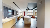 This thoughtfully designed single-floor amenity lounge delivers a vibrant, urban atmosphere through a blend of industrial and contemporary elements. Exposed ductwork and dark painted ceilings contrast with white walls, while track lighting throughout highlights bold, colorful artwork displayed across the space.

The lounge is organized into distinct yet open zones. A game room anchors one end, featuring a billiards table, a bar-height counter with teal stools, and eclectic seating arranged atop a vivid abstract area rug. Adjacent to this, a generous living area offers a large tufted sectional sofa, accent chairs, and a wall-mounted television—ideal for gathering and relaxation.

A sleek fireplace with dark tile surround serves as a striking focal point in a quieter seating nook, framed by large windows that flood the space with natural light and offer views of an adjacent outdoor terrace and urban surroundings. A sliding glass door provides direct access to the terrace.

The wet bar area features warm gold cabinetry, a mosaic tile backsplash, an under-counter refrigerator, pendant lighting, and a substantial marble-topped island with bar seating. Wood-look flooring flows throughout, unifying the space with warmth and continuity.