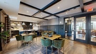Located at 2050 Stanford Ln in Newport News, VA, this single-floor community space is thoughtfully designed with a refined, upscale aesthetic that blends comfort with sophistication. The interior is unified by a deep navy and charcoal color palette, decorative wainscoting, and rich wood-look flooring laid in a herringbone pattern, creating a cohesive and polished atmosphere throughout.

The living area serves as a welcoming focal point, anchored by a sleek linear fireplace set within a built-in display wall. Plush seating, gold-accented round coffee tables, and a striking multi-arm brass chandelier overhead contribute to an inviting, lounge-like ambiance. Large sliding glass doors flood the space with natural light and provide direct access to an outdoor area.

The dining zone features multiple wood-top tables surrounded by tufted green velvet chairs, ideal for group gatherings. Exposed dark ceiling beams add architectural character and visual depth to the open layout.

The kitchen is a standout feature, outfitted with floor-to-ceiling navy cabinetry with brass hardware, a white subway tile backsplash, a generously sized waterfall-edge island with an integrated sink, and pendant lighting. Under-cabinet accent lighting and ample counter space make it both functional and visually impressive, well-suited for community events and catered functions.