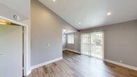 Located at 10990 Del Norte St in Ventura, California, this efficiently designed single-floor unit offers a thoughtfully arranged layout encompassing a living area, dining room, kitchen, bedroom, bathroom, and closet. The living and dining areas are characterized by striking vaulted ceilings that amplify the sense of space, complemented by light gray walls, recessed lighting, and warm wood-look flooring throughout. A sliding glass door floods the living area with natural light and provides views of lush outdoor greenery. The dining space features a decorative pendant light fixture, adding a refined touch. The kitchen is well-equipped with white shaker cabinetry, granite-style countertops, a stainless steel sink, and stainless steel appliances including a refrigerator and range. The bedroom is generously proportioned with mirrored sliding closet doors, large windows overlooking mature trees, and the same consistent wood-look flooring. An adjacent area leads to a bathroom featuring a full tub/shower combination with a curtain rod and a frosted window for natural light. White vanity cabinetry and a well-lit mirror complete the bathroom. The unit presents a clean, modern aesthetic with a cohesive neutral palette throughout.