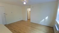 This single-floor space offers a clean, straightforward layout designed for comfortable everyday living. The main living area is finished with warm-toned hardwood floors that run throughout, lending a sense of continuity and warmth to the space. Crisp white walls and a neutral color palette keep the interiors feeling bright and open, while a ceiling fan with a light fixture provides both air circulation and overhead lighting.

Natural light filters into the living area, casting a warm glow across the hardwood floors and highlighting the simplicity of the space. A doorway connects the main room to an adjacent area, which appears to feature a closet with classic louvered bifold doors — a practical storage solution that adds a traditional touch. The transition between rooms is seamless, with consistent flooring and wall tones maintaining a cohesive aesthetic throughout.

The layout is efficient and uncluttered, offering a blank canvas that can be easily personalized. Electrical outlets are conveniently positioned along the baseboards, supporting flexible furniture arrangements. Overall, the space presents a tidy, well-maintained interior with timeless finishes that balance practicality with a welcoming ambiance.