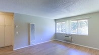 This single-story space is a straightforward, functional living area finished with light-toned hardwood-style flooring that runs throughout, lending warmth and easy maintenance to the interior. The walls are painted in a soft, neutral gray-blue tone, creating a calm and understated atmosphere. A textured popcorn ceiling adds a classic, utilitarian character to the room.

Natural light enters through a pair of windows fitted with sheer white curtains, offering views of mature trees outside and brightening the interior during the day. The windows are positioned along one wall, contributing to an open and airy feel within the space.

Climate control is addressed through two dedicated systems: a tall, wall-mounted vertical heating unit positioned along the main wall, and a through-the-wall air conditioning unit installed beneath the windows, providing year-round temperature management. White baseboards frame the perimeter of the room, offering a clean contrast against the gray-blue walls.

Overall, the space presents a clean, no-frills layout well-suited for practical, comfortable everyday living, with essential amenities already in place.