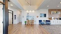 The Bend on Promenade leasing office in Rogers, Arkansas offers a thoughtfully designed, single-floor space that makes a strong first impression. The open-concept reception area greets visitors with warm wood-look tile flooring, a dramatic tray ceiling with elevated height, and a striking sculptural driftwood reception desk positioned beneath a sleek pendant light fixture. The community's branded signage is prominently displayed on a crisp white accent wall, setting a polished, modern tone throughout.

Adjacent to the entry, a spacious lounge area transitions to plush carpet and is bathed in natural light from multiple windows with views of the surrounding landscape. This area is furnished with a mix of cognac leather and navy accent seating, a geometric metallic coffee table, and curated wall art, creating a sophisticated yet welcoming atmosphere. A dedicated refreshment station features white cabinetry topped with a warm brick tile accent wall, a wall-mounted TV, and small appliances for resident and guest convenience.

A glass-panel private office or conference room is accessible from the main floor, offering transparency while maintaining separation. The overall design blends contemporary style with warm, residential touches, reflecting the character of the community it represents.