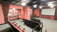 This single-floor private theater space is designed to deliver an authentic cinematic experience. The room is dressed in warm terracotta-red walls accented by dark wood ceiling beams and rich burgundy draping curtains that line the perimeter, creating an intimate and dramatic ambiance reminiscent of a classic movie house. Rows of plush, dark upholstered theater seats with built-in cup holders are arranged in a tiered layout facing a large projection screen, ensuring clear sightlines from every position. Pendant lighting and wall-mounted sconces provide soft, atmospheric illumination throughout the space. A standout feature is the vintage-style popcorn cart stationed near the entrance, adding a charming and nostalgic touch. Movie-themed framed artwork decorates the walls, reinforcing the cinematic theme. At the rear of the room, high-top tables and chairs offer an alternative viewing area for those who prefer a more casual seating arrangement. A ceiling-mounted projector and security camera are also visible, highlighting the room's functional infrastructure. The carpeted flooring and enclosed layout contribute to excellent acoustics, making this space well-suited for private screenings and entertainment gatherings.