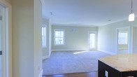 This single-floor space features an open layout that combines a living area with an adjacent kitchen space. The living area is finished with plush carpeted flooring, while the kitchen transition area features hardwood-style flooring, creating a clear visual distinction between the two zones. Soft, neutral green-toned walls are complemented by white crown molding and clean white baseboards, lending the space a classic yet understated aesthetic. Multiple windows along the exterior walls allow generous amounts of natural light to pour in, enhancing the bright and airy atmosphere throughout the room. A glass-paneled door provides access to an outdoor area, further amplifying the sense of openness and connectivity to the exterior. A visible HVAC vent on the wall ensures climate control functionality. The kitchen area, partially visible, features dark cabinetry paired with light granite countertops, offering a pleasant contrast to the overall neutral palette. The space presents a clean, move-in ready condition with a simple, functional design suitable for comfortable everyday living.