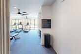 This fitness center in Pittsburgh, PA (15205) offers a thoughtfully designed, single-floor workout space with a generous open layout. The facility is divided into distinct functional zones, providing ample room for both cardio and strength training activities. The cardio area features multiple treadmills and elliptical machines positioned along large floor-to-ceiling windows, flooding the space with natural light and offering views of the surrounding greenery. Overhead, large modern ceiling fans provide comfortable airflow throughout. The strength training area is well-appointed with a multi-station cable machine, a full rack of kettlebells and dumbbells, and additional resistance equipment. A large wall mirror enhances the sense of space and supports proper form during workouts. The flooring transitions between blue carpet and durable grey fitness flooring depending on the zone. A slatted wood accent wall adds a warm, contemporary architectural touch near the entrance, which also includes built-in open shelving for storage. A bold black textured tile accent wall anchors a hydration station and storage console, adding a striking design element. Recessed lighting and posted fitness center policies complete this clean, modern, and fully functional facility.