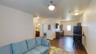 Located at 5629 Elysian Road in Billings, Montana, this single-story home features an open-concept layout that connects the main living spaces in a practical and functional arrangement. The living area is anchored by warm hardwood-style flooring that extends throughout the unit, providing a cohesive look across the space. A pendant light fixture hangs from the ceiling, adding a decorative touch above the seating area.

The kitchen runs along one wall and is equipped with dark cabinetry, a sink positioned beneath a window that allows natural light to filter in, and a black refrigerator near the entry door. The adjacent dining area accommodates a small table, making the kitchen and dining zone efficient for everyday use.

An electrical panel is visibly integrated into the wall between the living and kitchen areas. Beyond the main living space, a hallway leads to the bedroom and a bathroom, both of which are partially visible from the main area. The bathroom features wood-toned cabinetry and a vanity. The home's neutral wall tones and consistent flooring throughout create a clean, cohesive atmosphere well-suited for comfortable everyday living.