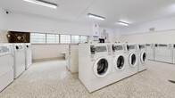 Located at 33 Pears Ave in Toronto's Annex neighbourhood, this shared laundry facility offers a well-organized and functional space for residents. The room is finished with durable terrazzo flooring and white tile wainscoting along the walls, contributing to a clean and easy-to-maintain environment. Fluorescent strip lighting mounted to the white ceiling provides bright, even illumination throughout.

The facility is equipped with multiple front-loading washers and top-loading washing machines, all featuring digital coin-operated controls, alongside a row of top-loading dryers. A utility sink is available for hand-washing or pre-treatment needs, and plumbing hookups along one wall indicate additional machine capacity. A wall-mounted clock helps residents manage their laundry time efficiently.

A row of windows along one wall allows natural light to supplement the overhead lighting, adding to the open and airy feel of the space. A small bookshelf stocked with reading material provides a thoughtful amenity for residents waiting during their laundry cycle. An information board and posted notices keep users informed of facility guidelines. Safety features include a fire extinguisher and clearly marked signage. The space is practical, well-maintained, and designed to serve the everyday laundry needs of building residents effectively.