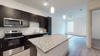 Located at 6001 S Congress Ave in Austin, Texas, this single-floor residence offers a thoughtfully designed open-concept layout with contemporary finishes. The kitchen serves as the centerpiece of the living area, showcasing rich dark cabinetry, a mosaic tile backsplash, and a large granite-topped island perfect for dining or meal prep. Stainless steel appliances — including a refrigerator, range, and microwave — complement the sleek aesthetic, while light wood-look flooring and pendant lighting add warmth and style. The open flow connects the kitchen seamlessly to the living area, which features recessed lighting, a ceiling fan, and windows that invite natural light. An in-unit stacked washer and dryer is conveniently tucked away near the main living space. The bedroom areas include a carpeted secondary room with a window, and a primary suite with light-toned wood flooring, a ceiling fan, and access to natural light. The primary bathroom is well-appointed with a granite-topped dark vanity, tile flooring, a glass-enclosed walk-in shower with full tile surround, and direct access to a spacious walk-in closet with built-in shelving. The overall ambiance is clean, bright, and modern throughout.