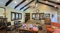 This single-story space showcases a striking vaulted ceiling accented by bold, dark-stained exposed wooden beams that draw the eye upward and lend a rustic, lodge-inspired character to the interior. A statement antler chandelier serves as a dramatic focal point, complementing the space's warm, nature-inspired aesthetic. The main living area is generously proportioned, offering an open and airy feel enhanced by recessed lighting and multiple windows fitted with wood-trimmed blinds that allow controlled natural light.

The seating area centers around a tufted gray sofa paired with classic leather armchairs featuring turned wooden details, all arranged around a substantial wooden coffee table set atop a richly patterned area rug. A large dark wood bookcase provides both storage and display space, contributing to the room's traditional, well-appointed feel. A decorative wall clock and framed artwork add personalized touches throughout.

French doors visible along one wall introduce additional natural light while connecting the interior to an adjacent space. The wood-look flooring throughout ties the design together, reinforcing the warm, inviting atmosphere that defines this traditionally styled, character-rich space.