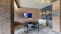 This covered outdoor amenity space offers a well-appointed gathering area defined by its striking exposed brick architecture and open-air design. Large brick columns frame the space throughout, lending a warm, industrial character that pairs naturally with the stone tile flooring underfoot. Decorative globe lights are strung along the ceiling beams, creating an inviting ambiance during both daytime and evening use.

The space is divided into distinct functional zones. A dining area centers around a dark rectangular table with stylish wire-back chairs, positioned beneath a wall-mounted flat-screen television set against a full exposed brick wall. Adjacent to the dining area, a built-in wet bar features white cabinetry, a stainless steel under-counter refrigerator, and a dramatic backdrop of patterned dark tile, illuminated by elegant gold pendant lights.

A generous lounge area offers comfortable outdoor seating with upholstered chairs and sofas arranged around the brick columns, ideal for relaxed gatherings. Black wrought-iron fencing and gated openings connect the space directly to a pool courtyard beyond, allowing natural light to pour in and providing open views of the surrounding community. The overall ambiance is social and polished, blending rugged brick textures with refined finishes to create a versatile, resort-style outdoor retreat.