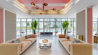 Located in Miami's dynamic NW corridor, this generously proportioned amenity lounge is a masterclass in contemporary tropical design. The single-floor space is defined by soaring floor-to-ceiling glass walls that frame sweeping views of an outdoor pool and surrounding urban landscape, flooding the interior with abundant natural light throughout the day.

The lounge is divided into distinct yet connected zones. A welcoming seating area near the entrance features a coffered tray ceiling with bold coral and white stripes, anchored by a sculptural mid-century-inspired brass chandelier. Plush, blush-toned sofas flank a marble-topped gold coffee table, creating an elegant yet relaxed gathering spot. Towering tropical plants punctuate the space, reinforcing the Miami aesthetic.

A central co-working and social bar area showcases a vibrant mosaic tile accent wall in a kaleidoscope of colors, with pendant lighting above a light wood counter and barstool seating — ideal for casual work or conversation. Additional lounge seating with warm-toned wood chairs adds a lively, social character.

A media lounge anchors the far end of the space, featuring a bold yellow graphic mural wall, large-screen TV, wraparound white sectional sofa, a tropical-motif area rug, and modern ring chandelier lighting. Arched architectural details with teal insets and gold sconces lend a polished, resort-inspired finish throughout.