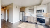 This single-floor space showcases a thoughtfully designed, contemporary interior with a clean, modern aesthetic throughout. The kitchen serves as a central highlight, featuring a full suite of stainless steel appliances, including a refrigerator, dishwasher, gas range, and built-in microwave. Cabinetry is finished in a two-tone palette, combining crisp white upper cabinets with warm wood-toned accents. A white subway tile backsplash adds texture, while a matte black faucet over an undermount sink provides a refined contrast. Dark hardwood-style flooring runs throughout the space, grounding the light-toned walls and cabinetry. A large window in the kitchen fills the space with natural light and offers open views of the surrounding cityscape, enhancing the airy atmosphere. The bathroom is finished with a light wood vanity topped with white quartz countertop, a black-framed mirror, matte black fixtures, and a tiled walk-in shower. A dedicated in-unit laundry closet houses a stacked Whirlpool washer and dryer, adding practical convenience. Overall, the space presents a cohesive, modern design with quality finishes and functional amenities.