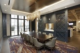 This sophisticated single-floor gathering space blends upscale design with functional versatility, making it ideal for private dining, corporate meetings, or intimate events. At the heart of the room sits a substantial dark-wood conference table surrounded by plush upholstered chairs, all anchored by a richly patterned area rug in deep blues, purples, and golds that sets a dramatic tone underfoot.

Overhead, two cascading gold chain chandeliers serve as the room's crown jewels, casting a warm, ambient glow against a dark ceiling that adds depth and intimacy. A boldly textured feature wall, dressed in an eye-catching ogee-patterned wallcovering in gold and charcoal tones, is illuminated by recessed accent lighting to create a striking focal point.

The space is framed by floor-to-ceiling dark-framed glass doors that invite abundant natural light and offer views of an outdoor terrace, seamlessly connecting the interior to the exterior. Deep navy sliding barn-style doors with latticed glass panels provide an elegant partition, concealing a built-in service area with a pass-through window. Along one wall, a sleek built-in credenza with a marble-style countertop and gold hardware is paired with a mounted flat-screen display, adding a practical yet refined touch. The overall ambiance is refined, dramatic, and welcoming.