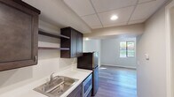 Located at 12730 Monte Vista in Poway, Unit #225 is a single-floor unit thoughtfully designed to balance functionality with comfort. The space features light-toned, wood-look plank flooring throughout the main living area, neutral gray walls, and generous natural light streaming in through a window that offers views of surrounding greenery.

The kitchenette is well-appointed with dark espresso-stained cabinetry, open display shelving, a white countertop, a stainless steel double-basin sink, a built-in microwave, and a full-sized refrigerator. A drop-panel ceiling with recessed lighting gives the kitchen area a clean, modern finish. The kitchen flows seamlessly into the open living area, creating an efficient and cohesive layout.

The bathroom is designed with accessibility in mind, featuring grab bars positioned near the toilet and throughout the walk-in shower. The shower includes a built-in bench seat, white subway tile with a decorative mosaic accent border, and a recessed shelf for added convenience. Light tile flooring completes the space.

The main living area is open and versatile, with clean lines, ample wall space, and a bright window that enhances the airy ambiance. Overall, the unit presents a practical and modern living environment suited for a range of needs.