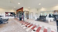 Located at 7298 Kensington Way in McCordsville, Indiana, this fitness center offers a generously sized, single-floor exercise space designed to accommodate a comprehensive range of workout needs. The room is defined by a striking combination of deep crimson accent walls and crisp white finishes, creating an energizing yet clean atmosphere throughout.

The space is thoughtfully organized into distinct workout zones. The cardio area features multiple treadmills, elliptical machines, stationary bikes, spin cycles, and a stair climber, all positioned to take advantage of the large windows that line the walls and invite abundant natural light and outdoor views of the surrounding greenery. A dedicated strength training section houses a cable and multi-function weight machine, a rack of dumbbells, and a weight bench, supported by built-in shelving for storage.

Recessed ceiling lighting supplements the natural light, ensuring even illumination across the entire floor. A ceiling fan aids in ventilation and comfort during intense workouts. Full-length mirrors along one wall enhance the sense of openness and allow users to monitor their form. The flooring features a distinctive patterned tile with red, gray, and beige tones, complemented by rubber mat sections in designated lifting areas. The overall layout is open and uncluttered, making this fitness center both functional and inviting.