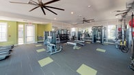 This single-floor fitness center is thoughtfully designed to accommodate a wide range of workout styles and fitness goals. The space is divided into distinct functional zones, each outfitted with professional-grade equipment. The strength training area features a full rack of dumbbells, multiple selectorized cable machines, and weight benches, all reflected in floor-to-ceiling mirrors that enhance the sense of openness. A dedicated cardio zone lines one wall with multiple treadmills and elliptical machines, positioned near windows that bring in natural light and views of the surrounding exterior. A functional training area adds versatility, housing a heavy bag, battle ropes, medicine balls in varying weights, plyo boxes, and a wall-mounted flat-screen television — ideal for high-intensity interval or circuit-style training. The flooring throughout is a dark commercial-grade carpet with chartreuse accent squares, providing both durability and visual interest. Oversized ceiling fans with wide blade spans circulate air efficiently across the main floor, while recessed lighting ensures consistent illumination. Chartreuse-painted accent walls in select areas add an energetic, modern touch to the overall aesthetic. The facility also includes a water station and direct exterior access, making it both functional and convenient.