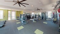 This single-floor fitness center is thoughtfully designed to accommodate a wide range of workout styles and fitness goals. The space is divided into distinct functional zones, each outfitted with professional-grade equipment. The strength training area features a full rack of dumbbells, multiple selectorized cable machines, and weight benches, all reflected in floor-to-ceiling mirrors that enhance the sense of openness. A dedicated cardio zone lines one wall with multiple treadmills and elliptical machines, positioned near windows that bring in natural light and views of the surrounding exterior. A functional training area adds versatility, housing a heavy bag, battle ropes, medicine balls in varying weights, plyo boxes, and a wall-mounted flat-screen television — ideal for high-intensity interval or circuit-style training. The flooring throughout is a dark commercial-grade carpet with chartreuse accent squares, providing both durability and visual interest. Oversized ceiling fans with wide blade spans circulate air efficiently across the main floor, while recessed lighting ensures consistent illumination. Chartreuse-painted accent walls in select areas add an energetic, modern touch to the overall aesthetic. The facility also includes a water station and direct exterior access, making it both functional and convenient.