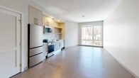 This single-floor unit offers a thoughtfully designed open-concept layout that seamlessly integrates the living and kitchen areas. Light-toned hardwood-style flooring flows throughout the main living space, contributing to a bright and cohesive aesthetic. A large picture window in the living area floods the space with natural light and provides views of surrounding greenery, enhancing the overall sense of openness.

The kitchen is efficiently designed and well-appointed, featuring light wood cabinetry with sleek bar-pull hardware, a white tile backsplash, and a full suite of stainless steel appliances including a refrigerator, range, over-the-range microwave, and dishwasher. A built-in open shelf adds practical display or storage space, while the undermount sink with a modern faucet completes the functional layout.

A hallway connects the living area to additional rooms, including a closet with louvered doors offering convenient storage access. The bathroom presents a clean, minimalist design with dark tile flooring, a wall-mounted white vanity with an integrated sink, contemporary chrome fixtures, and a large frameless mirror. Neutral wall tones throughout the unit create a calm, versatile backdrop, making the space move-in ready and easy to personalize.