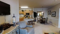 This single-floor residence showcases an open-concept design that integrates the kitchen, dining, and living areas into one cohesive, flowing space. The kitchen is outfitted with light maple cabinetry, granite countertops, and black appliances, including a built-in microwave and range. A central island with bar stool seating provides additional workspace and casual dining options. Crown molding adds a refined architectural touch throughout the space. The dining area accommodates a round table with seating for multiple guests and is accented by a pendant light fixture and a decorative mirror that enhances the sense of depth in the room. A tall indoor plant adds a natural element to the corner. The living area features plush, striped upholstered seating and is anchored by neutral-toned carpeting that runs throughout the common spaces. Warm, ambient lighting from both overhead fixtures and pendant lights contributes to the inviting atmosphere. The layout is well-suited for everyday living and entertaining, with each zone clearly defined yet openly connected.