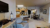This single-floor residence showcases an open-concept design that integrates the kitchen, dining, and living areas into one cohesive, flowing space. The kitchen is outfitted with light maple cabinetry, granite countertops, and black appliances, including a built-in microwave and range. A central island with bar stool seating provides additional workspace and casual dining options. Crown molding adds a refined architectural touch throughout the space. The dining area accommodates a round table with seating for multiple guests and is accented by a pendant light fixture and a decorative mirror that enhances the sense of depth in the room. A tall indoor plant adds a natural element to the corner. The living area features plush, striped upholstered seating and is anchored by neutral-toned carpeting that runs throughout the common spaces. Warm, ambient lighting from both overhead fixtures and pendant lights contributes to the inviting atmosphere. The layout is well-suited for everyday living and entertaining, with each zone clearly defined yet openly connected.