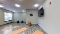 This single-floor wellness studio offers a serene and functional environment designed for yoga and light fitness activities. The room features smooth hardwood-style flooring in a neutral gray-brown tone, providing a clean and durable surface ideal for exercise. The walls are painted in a soft, muted gray, contributing to a calm and focused atmosphere throughout the space.

A decorative black lotus flower decal adorns one wall, serving as a mindful focal point that reinforces the studio's wellness-oriented theme. A large flat-screen TV is mounted on the adjacent wall, offering the ability to follow guided workout sessions or classes. Several peach-toned yoga mats are laid out across the floor, ready for use, while two stability balls are positioned toward the back of the room, adding versatility for fitness routines.

A woven basket near the back wall provides practical storage for additional rolled yoga mats or accessories. The space is well-lit by flush-mounted ceiling lights that cast an even, bright glow throughout, enhancing visibility without disrupting the tranquil ambiance. The overall design balances functionality with a peaceful, minimalist aesthetic.