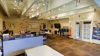 This single-floor creative studio or classroom is thoughtfully designed to support artistic and educational activities. The space features vaulted ceilings fitted with adjustable track lighting, providing customizable, well-distributed illumination across the entire room. The warm-toned walls are decorated with colorful hanging ornaments and student artwork, contributing to an engaging and lively atmosphere.

The layout is open and flexible, with multiple worktables arranged to accommodate group activities or individual projects. Along one side, a dedicated supply station is stocked with art materials including markers, paints, and organizers, keeping tools accessible and well-organized. A large whiteboard mounted on the wall serves as a focal point for instruction, accompanied by a wall-mounted flat-screen television for multimedia presentations.

Stacked chairs are neatly stored near the walls, suggesting the space can be reconfigured for different group sizes and activities. A door on the far wall provides additional access and allows natural light to filter into the room. The durable patterned carpet flooring adds a functional, low-maintenance quality suitable for high-traffic creative use. Overall, the space exudes a welcoming, activity-focused environment well-suited for classes, workshops, or creative programs.