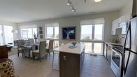 This single-floor coastal property offers an inviting open-concept layout designed to maximize its stunning waterfront surroundings. The kitchen and dining area flow together effortlessly, anchored by a sleek central island with a crisp white countertop and dark wood cabinetry, providing both functional workspace and casual seating. A full bank of large windows wraps the living and dining spaces, flooding the interior with natural light and framing unobstructed views of the water and shoreline beyond.

The dining area is furnished with woven rattan chairs and a wood table, lending a relaxed, coastal aesthetic that complements the neutral palette of soft whites and warm grays throughout. Nautical-inspired décor, including driftwood accents and ocean-themed artwork, reinforces the beachside character of the space. Light wood-toned flooring runs continuously through the open area, enhancing the airy feel.

Overhead, track lighting provides adjustable illumination to highlight key areas of the kitchen and living space. Roller shades on each window allow for privacy and light control without sacrificing the expansive views. The overall ambiance is calm, bright, and beach-inspired — a well-appointed space that brings the coastal landscape indoors.