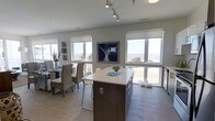 This single-floor coastal property offers an inviting open-concept layout designed to maximize its stunning waterfront surroundings. The kitchen and dining area flow together effortlessly, anchored by a sleek central island with a crisp white countertop and dark wood cabinetry, providing both functional workspace and casual seating. A full bank of large windows wraps the living and dining spaces, flooding the interior with natural light and framing unobstructed views of the water and shoreline beyond.

The dining area is furnished with woven rattan chairs and a wood table, lending a relaxed, coastal aesthetic that complements the neutral palette of soft whites and warm grays throughout. Nautical-inspired décor, including driftwood accents and ocean-themed artwork, reinforces the beachside character of the space. Light wood-toned flooring runs continuously through the open area, enhancing the airy feel.

Overhead, track lighting provides adjustable illumination to highlight key areas of the kitchen and living space. Roller shades on each window allow for privacy and light control without sacrificing the expansive views. The overall ambiance is calm, bright, and beach-inspired — a well-appointed space that brings the coastal landscape indoors.