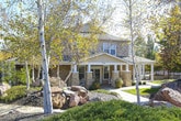 This single-story residence is situated within a thoughtfully landscaped community that immediately impresses with its natural surroundings. Winding concrete walkways guide visitors through a lush environment filled with mature deciduous and evergreen trees, neatly trimmed shrubs, ornamental grasses, and well-maintained mulched garden beds. The grounds offer a serene, park-like ambiance bathed in natural sunlight, creating an inviting approach to the home.

The exterior of the residence features classic horizontal lap siding in a warm neutral tone, complemented by white trim detailing that adds a clean, traditional aesthetic. A wall-mounted lantern-style light fixture accents the entryway, providing both functional illumination and architectural charm. The solid six-panel entry door, finished in a warm brown tone, offers a sturdy and straightforward entrance to the home.

The community setting enhances the overall appeal, with neighboring buildings of similar architectural style visible through the tree canopy, suggesting a cohesive and well-maintained residential environment. The combination of mature landscaping, tree-lined pathways, and classic exterior finishes gives this property a comfortable, established character that balances privacy with a welcoming sense of community.