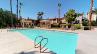 This single-level outdoor amenity space offers a resort-like atmosphere designed for relaxation and recreation. At its centerpiece is a generously sized swimming pool with crystal-clear turquoise water, accessible via stainless steel entry rails and surrounded by a spacious pool deck furnished with lounge chairs for sunbathing. Towering palm trees and mature shade trees frame the area, providing natural beauty and a sense of seclusion. Adjacent to the main pool is a circular spa, complete with seating and additional lounge chairs arranged along its perimeter, ideal for unwinding after a swim. A covered pergola area offers a shaded retreat, outfitted with a gas grill and outdoor dining table and chairs — perfect for casual gatherings and al fresco meals. An arched outdoor wet bar with a granite countertop and sink provides a convenient service station overlooking the pool area, adding a sophisticated touch to outdoor entertaining. The surrounding landscaping is lush and well-maintained, with trimmed hedges, flowering plants, and ornamental greenery that enhance the overall ambiance. The warm, Mediterranean-inspired architectural backdrop of stucco buildings with tiled rooflines completes the cohesive and inviting aesthetic of this outdoor community space.