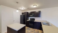 This single-story unit located at 3101 S Old 63 in Columbia, Missouri, offers a practical and cohesive layout designed for comfortable everyday living. The kitchen serves as a central highlight, showcasing dark espresso cabinetry, light countertops, and a full suite of black appliances including a refrigerator, electric range, built-in microwave, and dishwasher. A freestanding center island adds valuable prep space and visual contrast, while pendant lighting overhead and stone-look tile flooring complete the functional aesthetic. The kitchen flows openly into the living area, which features neutral carpet, a ceiling fan, and access to natural light through windows and a glass-paneled entry door. Bedrooms are carpeted and maintain the neutral color palette, each offering closet storage with built-in shelving. Two bathrooms echo the kitchen's design language, featuring dark vanity cabinetry, light countertops, large mirrors, and stone-look tile flooring. One bathroom includes a shower-tub combination with chrome fixtures. Throughout the unit, white panel doors, neutral walls, and consistent finishes create a clean, uniform ambiance.
