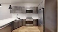 This thoughtfully designed one-bedroom, one-bathroom residence on Farmington Avenue in West Hartford, CT offers a well-appointed layout with modern finishes throughout. The kitchen serves as a focal point of the living area, showcasing dark shaker-style cabinetry with gold hardware, a white quartz peninsula with an undermount sink and matte black faucet, stainless steel appliances including a refrigerator, range, and built-in microwave, and a white tile backsplash. Stylish black pendant lights illuminate the space, complementing the warm hardwood-style flooring that extends through the main living area. The bedroom features plush carpeting, soft neutral wall tones, a double-door closet for ample storage, and a window with a roller shade that frames views of the surrounding West Hartford streetscape. The bathroom is finished with large-format floor tiles, a dark wood vanity with gold hardware, a white quartz countertop, marble-inspired wall tile, and a full soaking tub with a tiled surround. A dedicated in-unit laundry area with Samsung washer and dryer and built-in shelving above adds everyday convenience to this efficiently designed residence.