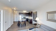 This well-appointed single-floor space offers a seamless open-concept layout that integrates the kitchen, dining, and living areas into one cohesive and functional environment. The kitchen is a standout feature, outfitted with rich dark espresso cabinetry that extends to the upper walls, providing ample storage. Stainless steel appliances — including a refrigerator, electric range, built-in microwave, and dishwasher — lend a sleek, modern aesthetic. Light-toned countertops contrast beautifully against the dark cabinetry, creating a striking visual balance. A kitchen island with bar-height seating for two serves as both a prep space and a casual dining spot, anchored by industrial-style adjustable stools. The dining and living areas flow naturally from the kitchen, unified by warm, wood-tone laminate flooring that runs throughout the space. The living area is bright and open, with neutral white walls that maximize the sense of airiness. Flush-mount ceiling lighting provides consistent illumination across the space. A private entry door is positioned conveniently near the kitchen, adding to the practical layout. Overall, the space combines contemporary design elements with functional living in a clean, modern aesthetic.