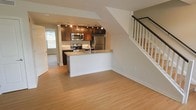 This two-story residence offers a thoughtfully designed open-concept main floor that seamlessly integrates the living and kitchen areas. The kitchen is a standout feature, showcasing rich dark wood cabinetry paired with granite countertops and a full suite of stainless steel appliances, including a refrigerator, microwave, and range. A peninsula with a built-in sink provides additional prep space and doubles as a casual dining or entertaining area. Track lighting illuminates the kitchen, highlighting the cabinetry and countertops effectively. The main living area is finished with light-toned hardwood flooring that extends throughout, creating a warm and cohesive aesthetic. A staircase with a dark handrail and white balusters leads to the upper floor, contributing a clean, modern architectural detail to the space. Natural light filters in through a window near the entry door, brightening the main level. The overall design balances functionality and style, with neutral wall tones and quality finishes that create a welcoming, move-in-ready environment across both floors.