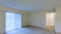 This single-floor space presents a clean, functional layout centered around a spacious main living area. The room is finished with wall-to-wall carpeting in a neutral tone, contributing to a quiet and comfortable atmosphere underfoot. Light-colored walls keep the interior feeling open and airy, while a textured ceiling adds a subtle architectural detail overhead.

A prominent sliding glass door dominates one wall of the living area, dressed with vertical blinds that allow for adjustable natural light and privacy control. The door provides direct access to an outdoor area, bringing in generous daylight that brightens the interior throughout the day.

Electrical outlets are positioned along the walls at practical intervals, offering flexibility for furniture arrangement and everyday use. A doorway visible toward one side of the room suggests additional spaces beyond the main living area, indicating a functional floor plan with clearly defined zones.

Overall, the space maintains a simple, no-frills aesthetic with a focus on practicality and ease of use. The neutral palette throughout provides a versatile backdrop suited to a variety of interior styles and furnishing choices.