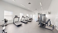 This fitness center offers a spacious, single-floor layout designed to accommodate a full range of workout activities. The space features a clean, minimalist aesthetic with crisp white walls, high ceilings, and linear LED lighting that creates a bright, energizing atmosphere throughout. Large windows line the walls, flooding the interior with natural light and providing views of the surrounding outdoor greenery.

The main workout area is generously proportioned, with a durable gray concrete-style floor that provides a functional surface for a variety of exercises. The cardio section is equipped with multiple treadmills and stationary cycling bikes, thoughtfully arranged to maximize space and usability. A dedicated strength training zone houses a multi-station cable machine, a power rack with an adjustable bench, and a full rack of dumbbells spanning a wide weight range, catering to users of all fitness levels.

A separate area accessible through glass-paneled doors provides additional functional training space, complete with wall-mounted resistance bands and supplementary equipment. The facility also features a bench near the entrance for convenience. With its open floor plan, abundant natural light, modern equipment, and thoughtful layout, this fitness center delivers a professional and inviting environment suited for comprehensive training.