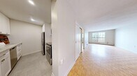 This thoughtfully designed one-bedroom, one-bathroom unit spans a single floor, offering a functional and comfortable layout. Warm parquet flooring flows consistently throughout the living room, bedroom, and hallways, lending a classic character to the interior. The living room is generously proportioned, bathed in natural light from a large window that offers views of the surrounding cityscape, and is equipped with baseboard heating for year-round comfort. The kitchen is efficiently laid out with crisp white cabinetry, subway tile backsplash, granite-look countertops, and stainless steel appliances including a refrigerator, range, and dishwasher — all finished with neutral tile flooring. The bedroom is a comfortable size, featuring a double sliding-door closet and a window that brings in additional natural light. The bathroom is clean and functional, appointed with a full soaker tub and shower combination, white subway tile surround, and chrome fixtures. Two hallways connect the spaces, providing a clear and logical flow throughout the unit. The overall palette is neutral and bright, with white walls and clean finishes creating a fresh, move-in-ready atmosphere.