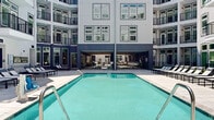 This expansive amenity complex is spread across two interconnected multi-story buildings, forming a well-appointed courtyard community designed for both relaxation and social engagement. At its centerpiece sits a large, resort-style swimming pool surrounded by a broad tiled pool deck lined with sleek lounge chairs, offering a bright, open-air atmosphere framed by the rising facades of the buildings above.

A spacious covered pavilion bridges the indoor-outdoor experience, featuring large floor-to-ceiling glass panels and wide-opening doors that look directly onto the pool area. Inside this sheltered lounge space, clean tile flooring, a built-in fireplace with stone detailing, plush sectional seating, a wall-mounted TV, and ceiling fans create a comfortable year-round gathering environment.

The exterior continues with a second courtyard area offering lush landscaped greenery, raised garden beds, and manicured turf — providing a quieter, garden-like retreat. Nearby, a dedicated outdoor entertainment zone includes multiple built-in grilling stations, a ping pong table, and bar-height seating with stone-clad table bases, all draped in ambient string lighting overhead.

The architecture throughout features a contemporary palette of white and deep navy siding, expansive grid-pane windows, and private balconies on upper floors, lending the complex a modern, polished aesthetic with a strong sense of community-oriented design.