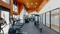Located at 901 E Van Buren St in Phoenix, AZ, this fitness center is a thoughtfully designed, single-floor facility comprising two dedicated exercise rooms. The space immediately impresses with its soaring double-height ceilings, accented by a bold, geometric orange upper-wall treatment that contrasts sharply with the crisp white lower walls, creating a dynamic and energizing atmosphere.

The main exercise room is generously proportioned and flooded with natural light through floor-to-ceiling glass panel walls and large windows, offering views of the surrounding outdoor pool area. The rubber-floored workout space is fully equipped with a wide array of strength and cardio equipment, including multi-station cable machines, a functional training rack, adjustable benches, a full dumbbell rack, treadmills, stair climbers, and elliptical machines. A ceiling fan provides additional airflow, complementing the recessed and pendant lighting overhead.

The second exercise room, separated by sleek black-framed glass sliding doors, offers a quieter, more focused training environment with hardwood-style flooring, making it ideal for stretching, functional training, or group sessions. Together, the two rooms provide a comprehensive and versatile fitness experience, enhanced by the bright Phoenix surroundings visible through the expansive windows throughout.