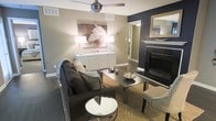 This single-floor space showcases a well-appointed living area with a refined contemporary design. The room is anchored by a plush dark sofa paired with a sleek rectangular coffee table, creating an inviting and functional seating arrangement. A circular mirrored accent table adds a touch of elegance to the space. The flooring features rich dark hardwood, which contrasts beautifully with the warm taupe and deep navy accent walls, lending depth and sophistication to the overall palette. A statement fireplace with dark stone surround serves as a dramatic focal point along one wall, while a large-framed equestrian artwork above a crisp white sideboard adds artistic character to the room. Twin table lamps flank the artwork, casting a warm ambient glow throughout the space. A ceiling fan with a brushed metal finish provides both style and practicality overhead. A patterned area rug grounds the seating area, adding texture and visual interest. A large mirror with a gold-toned frame reflects light and enhances the sense of openness. The cohesive design blends neutral tones with bold accents, resulting in a space that feels both elegant and comfortable.