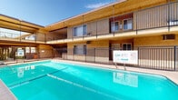 Located at 1206 W Fourth St in Ontario, California, this property features a centrally positioned outdoor swimming pool set within a courtyard-style layout. The pool is generously sized, with clear turquoise water and defined lane markings along the bottom. It is enclosed by a black iron safety fence and includes standard pool amenities such as metal entry rails and a life ring station mounted poolside.

The surrounding structure is two stories tall, finished in warm tan stucco with a flat-roofed design characteristic of mid-century California architecture. The upper level features open-air balconies with iron railings that overlook the pool area, while the ground floor units face the courtyard directly, many with sliding glass doors and decorative stonework accents. Rich burgundy-toned entry doors add a distinctive color contrast to the exterior facade.

Staircases at the corners of the building provide access between levels, and the layout creates an enclosed, semi-private atmosphere around the pool. The pool deck is finished in a light-toned concrete surface, offering ample space around the perimeter. The courtyard opens to clear southern California skies, allowing abundant natural sunlight to fill the space throughout the day. The overall setting is functional and communal, well-suited for outdoor recreation in the warm Ontario climate.