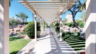 This beautifully designed outdoor space showcases a harmonious blend of structured architecture and natural landscaping. A long, pergola-covered walkway serves as the central feature, constructed from cream-colored columns and an open-slatted overhead grid that casts striking striped shadows across the concrete path below. The rhythmic pattern of light and shadow creates a visually engaging corridor that guides visitors through the grounds. Flanking the walkway are meticulously maintained hedgerows, manicured lawn sections, and large decorative boulders that lend a naturalistic, resort-like character to the environment. Tall, mature palm trees and shade trees rise throughout the landscape, adding vertical drama and a warm, sun-drenched ambiance consistent with a desert climate setting. Large ceramic and stone planters positioned along the pergola corridor contribute additional greenery and a refined aesthetic. Adjacent to the walkway, a tranquil pond with a central fountain serves as a focal point, its gently arching water jet surrounded by natural boulders and lush plantings. In the background, a Mediterranean-influenced building with a tile roof and neutral stucco exterior complements the overall design. The space exudes a serene, resort-quality atmosphere ideal for leisurely strolling and outdoor enjoyment.