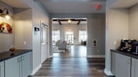 This single-floor community clubhouse blends modern farmhouse aesthetics with comfortable, functional design throughout its well-appointed spaces. The welcoming entry corridor features soaring vaulted ceilings with striking exposed dark wood beams, elegant cage-style pendant lanterns, and wide-plank hardwood-style flooring that flows seamlessly throughout the entire facility.

The main lounge area is a standout feature, highlighted by a dramatic floor-to-ceiling stone fireplace with a linear gas insert and a warm wood mantel, complemented by a wall-mounted TV above. Generous windows flood the seating area with natural light, offering views of the surrounding community and outdoor pool area. Exposed beam ceilings add architectural character and an open, airy ambiance.

A fully equipped kitchen serves the space with white shaker cabinetry, dark countertops, stainless steel appliances including a gas range with hood, refrigerator, and dishwasher, plus a center island with bar seating. A dedicated dining area adjacent to the kitchen features floor-to-ceiling windows overlooking the outdoor amenity area.

A billiards room provides additional recreation, while a separate intimate lounge nook offers a quieter retreat. The central hallway includes built-in cabinetry with countertops ideal for refreshment service. Restrooms are conveniently accessible throughout the space.