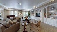 Located at 2290 Mackenzie Creek Rd in Chula Vista, CA, this refined single-floor community space is thoughtfully designed to offer both comfort and functionality across several distinct areas. The main lounge features plush leather seating, crown molding, and soft recessed lighting, anchored by a handsome grand piano that serves as a focal point. Built-in arched shelving with a centrally mounted television adds an elegant, classical architectural touch, while large windows invite natural light throughout. French doors connect the lounge seamlessly to the adjoining spaces, creating an open and welcoming flow. The activity and game room is generously sized, accommodating multiple round dining and card tables paired with cushioned chairs on rich hardwood flooring. Tall, multi-pane French doors line the walls, offering views of the lush outdoor greenery and filling the room with abundant natural light. A wall-mounted mirror enhances the sense of openness. The kitchen is practical and well-organized, featuring white raised-panel cabinetry, ample counter space with warm-toned laminate surfaces, tile flooring, and full appliances including a gas range and dishwasher. Crown molding is a consistent design element throughout, lending the entire space a polished and cohesive aesthetic well-suited for community gatherings and social activities.