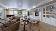 Located at 2290 Mackenzie Creek Rd in Chula Vista, CA, this refined single-floor community space is thoughtfully designed to offer both comfort and functionality across several distinct areas. The main lounge features plush leather seating, crown molding, and soft recessed lighting, anchored by a handsome grand piano that serves as a focal point. Built-in arched shelving with a centrally mounted television adds an elegant, classical architectural touch, while large windows invite natural light throughout. French doors connect the lounge seamlessly to the adjoining spaces, creating an open and welcoming flow. The activity and game room is generously sized, accommodating multiple round dining and card tables paired with cushioned chairs on rich hardwood flooring. Tall, multi-pane French doors line the walls, offering views of the lush outdoor greenery and filling the room with abundant natural light. A wall-mounted mirror enhances the sense of openness. The kitchen is practical and well-organized, featuring white raised-panel cabinetry, ample counter space with warm-toned laminate surfaces, tile flooring, and full appliances including a gas range and dishwasher. Crown molding is a consistent design element throughout, lending the entire space a polished and cohesive aesthetic well-suited for community gatherings and social activities.