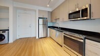 Located in Los Angeles, CA 90006, this modern single-floor unit offers a thoughtfully designed open-concept layout that seamlessly connects the kitchen and living area. The kitchen is well-appointed with a full suite of stainless steel appliances, including a refrigerator, electric range, built-in microwave, and dishwasher. Sleek dark countertops contrast elegantly with the warm, light-toned cabinetry that extends to the ceiling, providing ample storage. Track lighting overhead adds both functionality and a contemporary touch. Light wood-style flooring runs throughout the entire unit, creating a cohesive and clean aesthetic. The open living area is bathed in natural light, courtesy of large floor-to-ceiling windows fitted with horizontal blinds, offering views of the surrounding neighborhood. The bedroom is a bright, peaceful retreat featuring the same expansive windows that invite abundant daylight. The bathroom is finished with matching cabinetry, a dark countertop vanity, an integrated sink, a large mirror, and light wood flooring that ties into the rest of the unit. Crisp white walls and clean lines throughout contribute to the space's modern, minimalist character, making it a polished and functional urban residence.