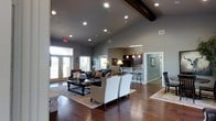This single-floor space makes a striking first impression with its soaring vaulted ceilings accented by rich, dark exposed wood beams. Recessed lighting is thoughtfully distributed throughout, ensuring the entire space is well-lit while complementing the warm, sophisticated ambiance. The open-concept living area is anchored by dark hardwood floors that run throughout, adding warmth and continuity to the design. The living room is generously proportioned, offering ample space for elegant seating arrangements, including plush upholstered chairs and a dark leather sofa centered around a substantial wooden coffee table, all set atop a decorative area rug. Large windows and French doors along one wall invite an abundance of natural light, creating a bright and welcoming atmosphere with views to the exterior. Flowing seamlessly from the living area, the kitchen is visible through an open pass-through, featuring dark cabinetry, pendant lighting, and a raised bar counter with bar stool seating — ideal for casual dining or entertaining. The cool gray wall tones throughout provide a modern, neutral backdrop that ties each area of the space together cohesively, resulting in an interior that is both functional and refined.
