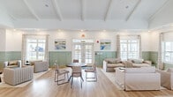 This single-floor clubhouse in North Augusta, SC, offers a generously sized, open-concept gathering space designed with a coastal-inspired aesthetic. The main lounge area is defined by soaring vaulted ceilings with exposed white beams, recessed lighting, and light-toned hardwood flooring that runs throughout the space. A standout white brick fireplace with a rustic wood mantel anchors one wall, flanked by French doors that invite natural light and outdoor views. The lower half of the walls is finished in sage-green wainscoting, a consistent design element that lends cohesion and charm across all areas.

Multiple seating arrangements accommodate both casual socializing and small group gatherings, while a round dining table with upholstered chairs provides a dedicated area for dining or meetings. A bar counter with a granite-speckled surface and bar stools serves as a functional focal point near the service corridor. A private office or conference room features the same wainscoting detail alongside a warm wood table, ideal for focused use. A long hallway connects the ancillary spaces, maintaining the light, airy palette throughout. Large windows dressed with plaid curtains allow natural light to fill the entire clubhouse.